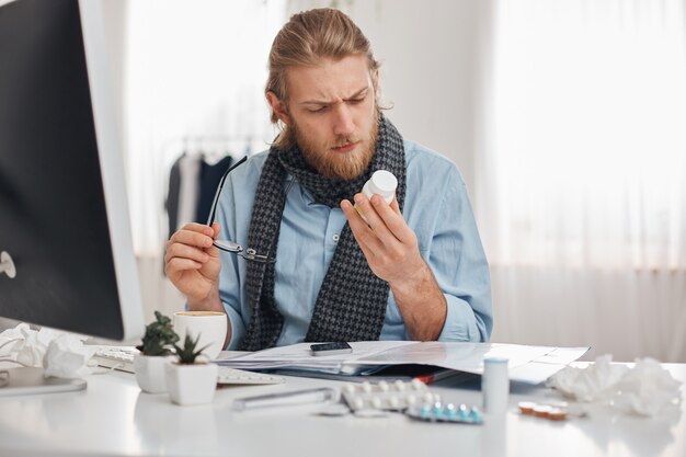 Больной бородатый мужской офисный работник в синей рубашке и шарфе с очками сосредоточены на чтении рецепта таблеток. Молодой менеджер с гриппом, сидит на рабочем месте в окружении лекарств, таблеток, витаминов