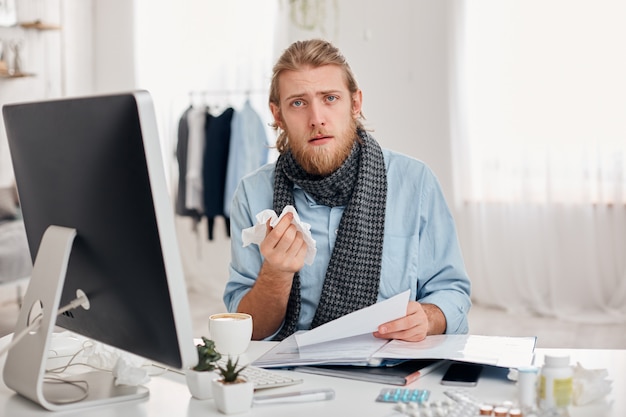 Ill bearded man sneezes, uses handkerchief, feels unwell, has flu. Sick male office worker has fever and tired expression, discusses working issues with colleagues. Illness and infection concept