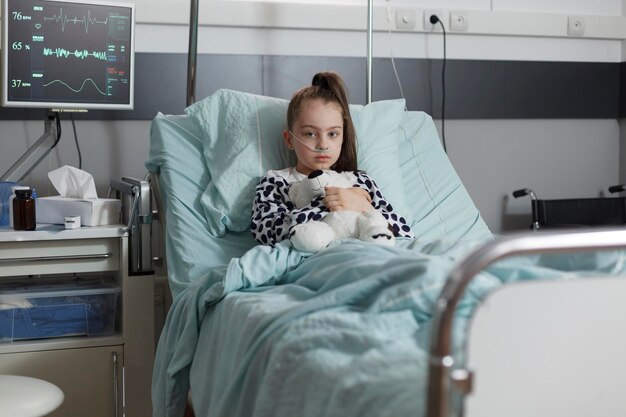 テディベアを持ちながら、子供の医療施設の患者のベッドで一人で休んでいる病気の子供。クマのぬいぐるみを持ちながら酸素チューブを装着して治療中の病気の少女。
