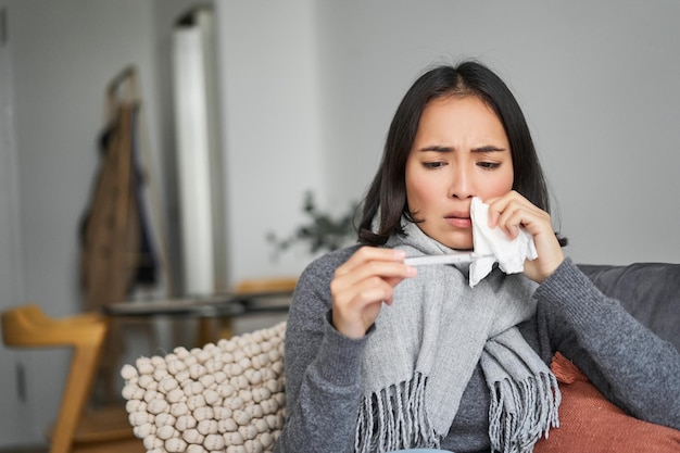 Free photo ill korean woman looking at thermometer with concerned face catching cold having fever having covid