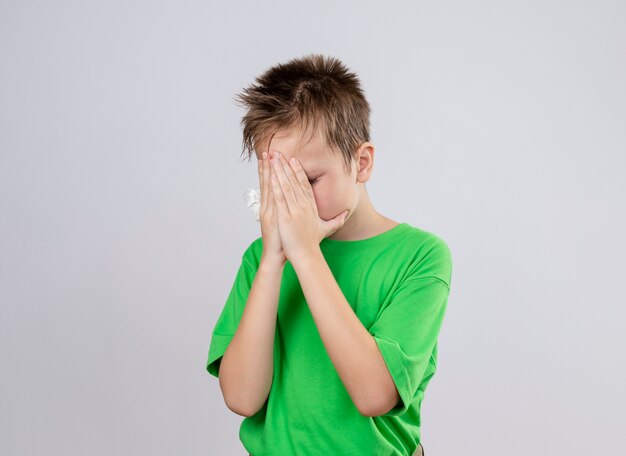 白い壁の上に立っている手で顔を覆う気分が悪い緑のTシャツの病気の小さな男の子