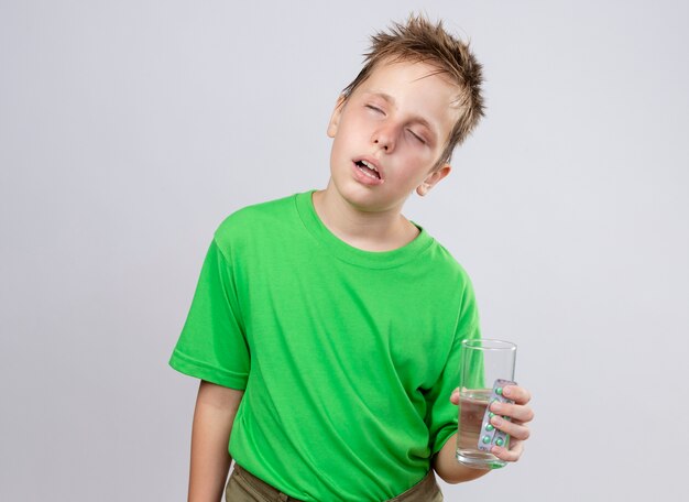 녹색 티셔츠에 아픈 어린 소년은 흰 벽에 감기 서서 고통받는 물과 알약 잔을 들고 몸이 좋지 않습니다.