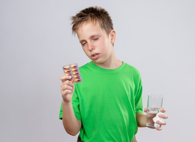 Ill little boy in green t-shirt feeling unwell holding glass of water and pills suffering from flu standing over white wall