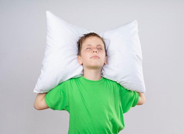 Ill little boy in green t-shirt feeling unwell holding pillow with closed eyes standing over white wall