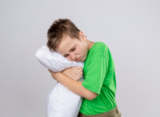白い壁の上に立っている寒さに苦しんで枕を抱き締める気分が悪い緑のTシャツの病気の小さな男の子
