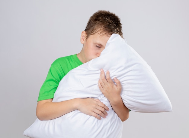 녹색 티셔츠에 아픈 어린 소년은 흰 벽 위에 서있는 닫힌 눈을 가진 몸이 안아주는 베개를 느끼고 있습니다.
