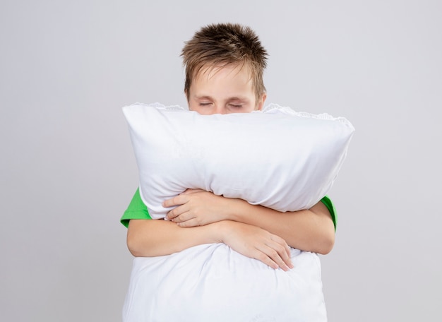 Free photo ill little boy in green t-shirt feeling unwell hugging pillow with closed eyes standing over white wall