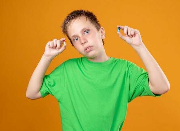 Ill little boy in green t-shirt feeling unwell showing pills in hands looking at camera being confused standing over orange background