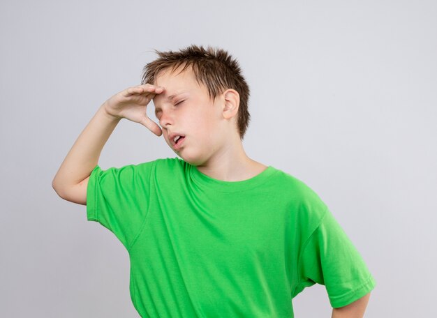 Больной маленький мальчик в зеленой футболке плохо себя чувствует, страдая от холода, стоя у белой стены
