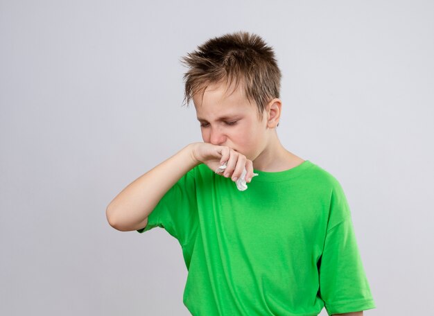 鼻水と白い壁の上に立っているインフルエンザに苦しんで気分が悪い緑のTシャツの病気の小さな男の子