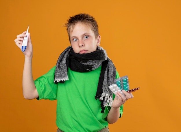 Ill little boy in green t-shirt and warm scarf around his neck holding pills showing thremometer looking  confusedstanding over orange wall