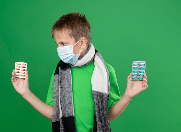 Free photo ill little boy in green t-shirt and warm scarf around his neckwearing facial protective mask holding pills looking confused trying to make choice standing over green background