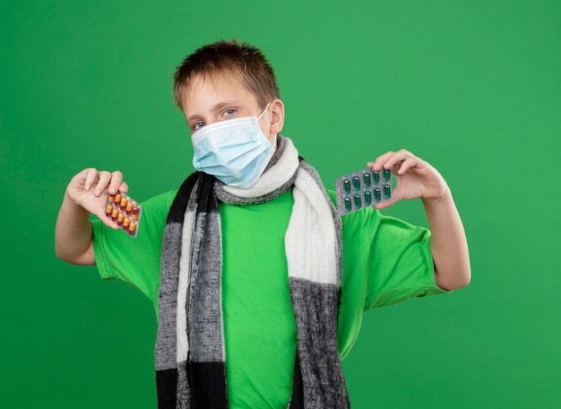 Ill little boy in green t-shirt and warm scarf around his neckwearing facial protective mask showing pills looking at camera standing over green background