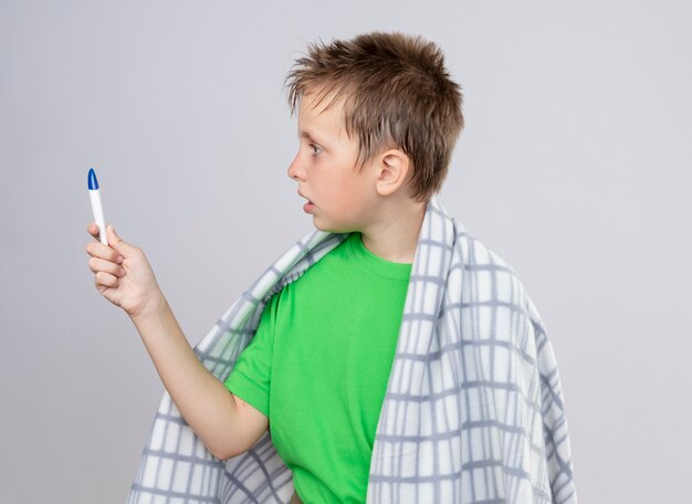 Ill little boy in green t-shirt wrapped in blanket feeling unwell holding thermometer looking at it amazed and worried standing over white wall