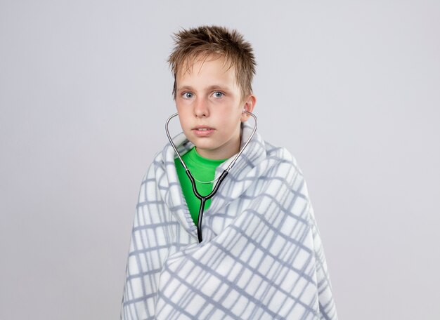 白い壁の上に立っている聴診器で気分が悪い毛布に包まれた緑のTシャツの病気の小さな男の子