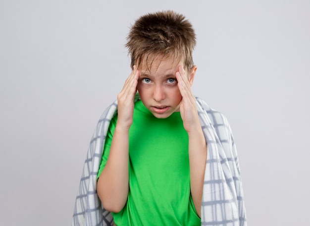 Бесплатное фото Больной маленький мальчик в зеленой футболке, завернутый в одеяло, чувствует недомогание, прикасаясь к вискам, чувствуя боль, стоя над белой стеной