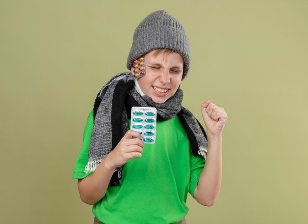 暖かいスカーフと帽子で緑のTシャツを着ている病気の小さな男の子は明るい壁の上に立っている拳を元気に握りしめながら笑顔のより良い丸薬を感じています