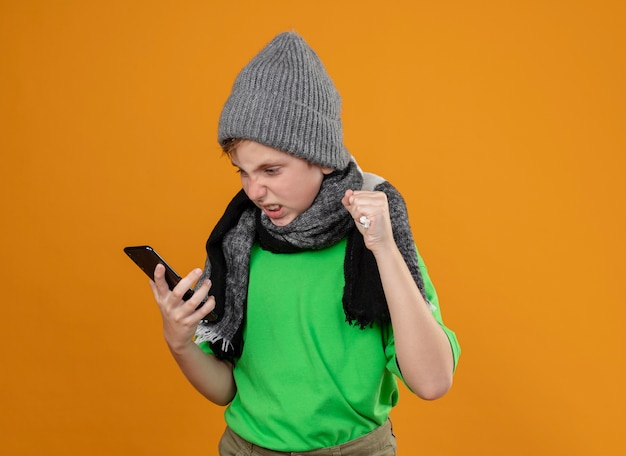 Ill little boy wearing green t-shirt in warm scarf and hat feeling unwell holding smartphone looking at it with angry expression standing over orange wall