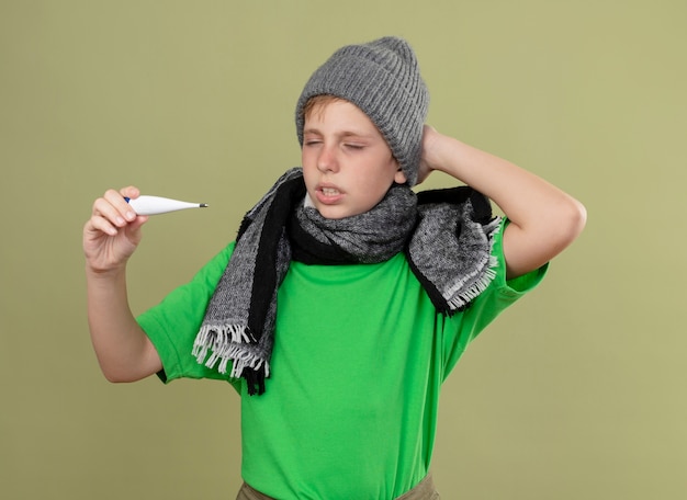 Free photo ill little boy wearing green t-shirt in warm scarf and hat feeling unwell holding thermometer looking at it confused standing over light wall