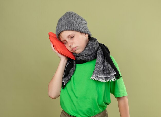 Ill little boy wearing green t-shirt in warm scarf and hat feeling unwell holding water bottle to keep warm leaning head on it standing over light wall