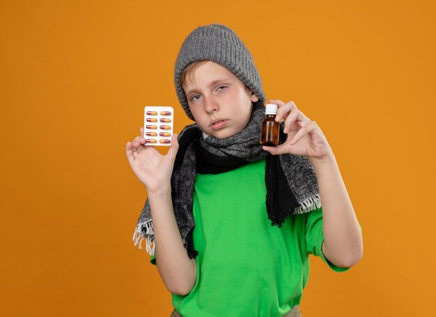 Больной маленький мальчик в зеленой футболке, теплом шарфе и шляпе чувствует себя нездоровым, показывая бутылку с лекарствами и таблетки, несчастный и больной, стоящий над оранжевой стеной