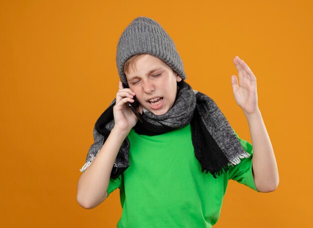 Ill little boy wearing green t-shirt in warm scarf and hat feeling unwell talking on mobile phone being displeased with annoyed expression standing over orange wall