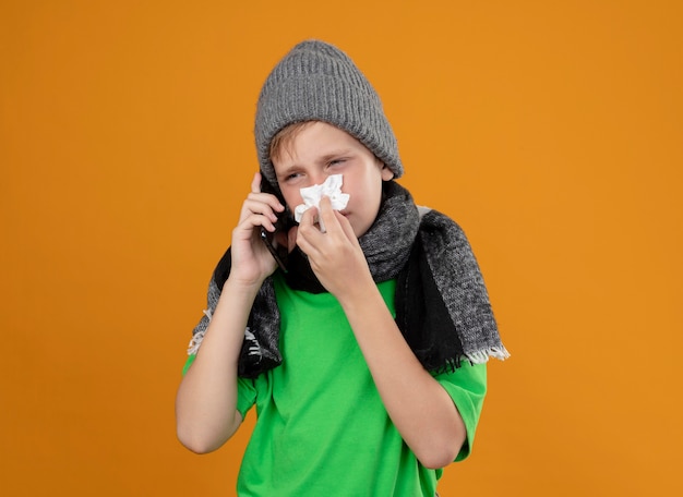 Ill little boy wearing green t-shirt in warm scarf and hat feeling unwell talking on mobile phone wiping nose with paper napkin suffering from cold standing over orange wall