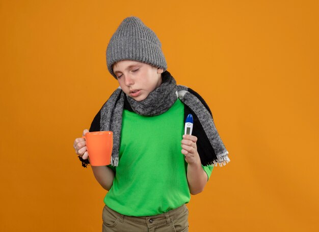 暖かいスカーフと帽子をかぶった緑色のTシャツを着て、熱いお茶と体温計を持った病気の少年