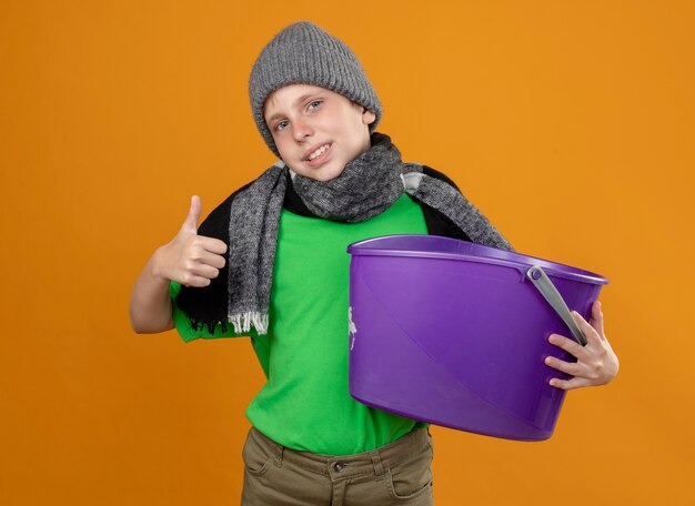 따뜻한 스카프와 모자에 녹색 티셔츠를 입고 아픈 어린 소년 오렌지 벽 위에 서있는 엄지 손가락을 보여주는 메스꺼움을 느끼는 쓰레기를 들고 모자