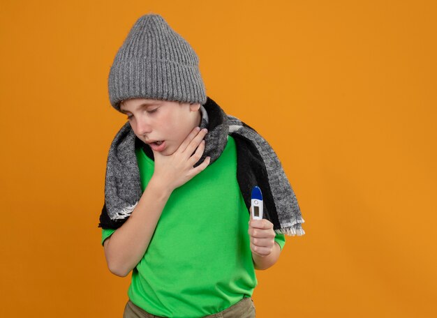 Больной маленький мальчик в зеленой футболке, теплом шарфе и шляпе, показывающий, что термометр плохо себя чувствует, больной и несчастный кашель стоит над оранжевой стеной