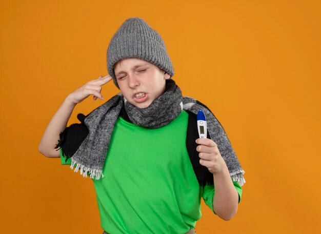 Ill little boy wearing green t-shirt in warm scarf and hat showing thermometer feeling unwell sick and unhappy making pistol gesture near temple standing over orange wall