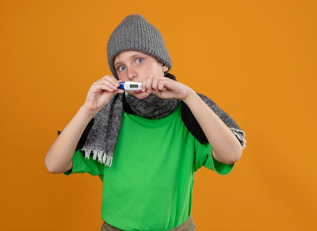 Ill little boy wearing green t-shirt in warm scarf and hat showing thermometer feeling unwell sick and unhappy standing over orange wall