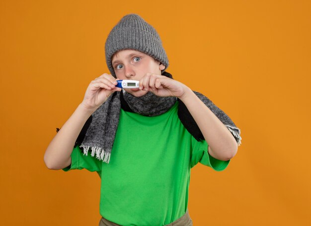 暖かいスカーフと帽子で緑のTシャツを着ている病気の少年は、温度計がオレンジ色の壁の上に立って気分が悪くて不幸に感じていることを示しています