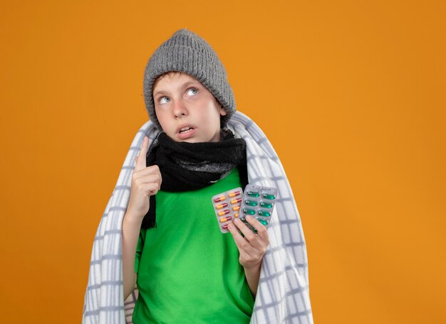 Ill little boy wearing warm hat and scarf wrapped in blanket suffering from fever holding pills feeling unwell looking up pointing with finger standing over orange wall