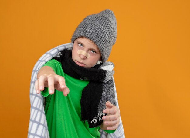 Ill little boy wearing warm hat and scarf wrapped in blanket with patch on his nose unhappy and sick looking  with arm out standing over orange wall