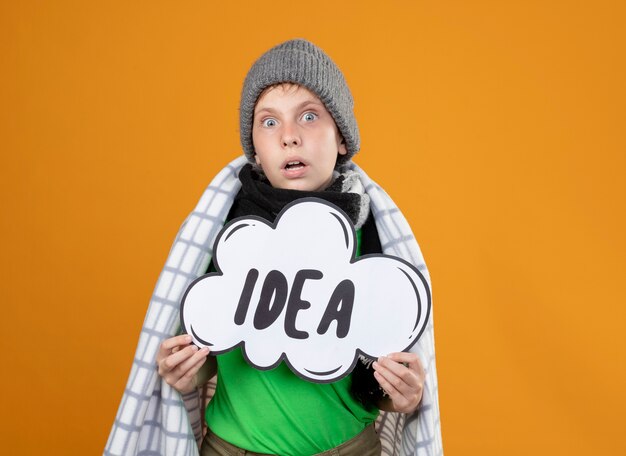 Ill little boy wearing warm hat and scarf wrapped in blanketshowing speech bubble sign with word idea looking  being surprised standing over orange wall