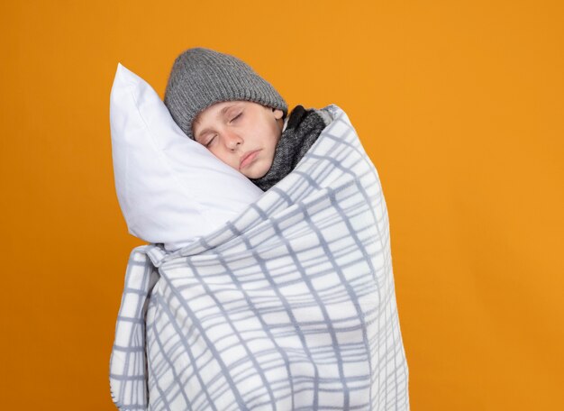 Ill little boy wearing warm hat wrapped in blanket suffering from fever holding pillow standing over orange wall