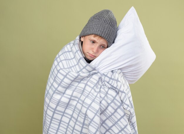 Ill little boyin warm hat wrapped in blanket holding pillow feeling unwell standing over light wall