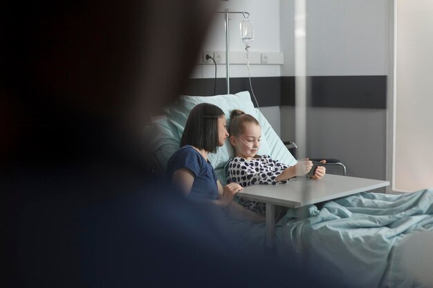 Ill little girl resting in hospital pediatrics ward while playing games on modern mobile phone. Mother sitting beside hospitalized daughter under treatment while watching cartoons on smartphone.