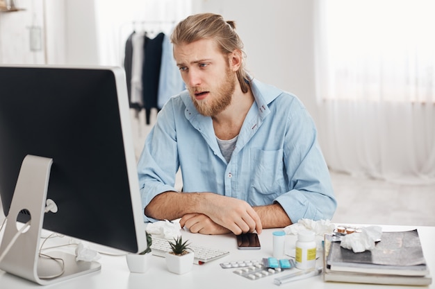 Foto gratuita uomo barbuto malato o malato vestito in camicia blu con espressione del viso stanco e sofferente, allergico, con problemi di salute. il giovane ha il naso che cola, si siede sul posto di lavoro con pillole e droghe