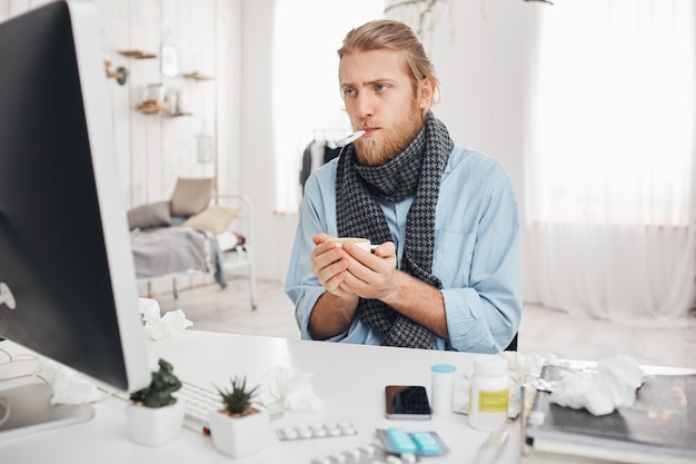Ill sick bearded male sits in front of computer screen with thermometer in mouth, measures temperature, holds a cup of hot drink in his hands. Sad young fair-haired man has a bad cold or flu