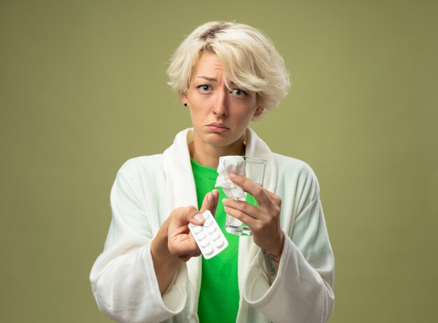 Ill woman with short hair feeling unwell holding glass of water and blister with pills looking  being unhappy with sad expression standing over light wall