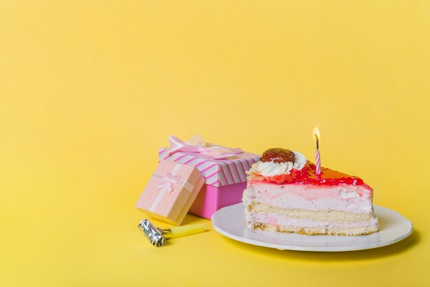Свеча свечи на ломтик торт с двумя подарочные коробки и участник вентилятор на желтом фоне
