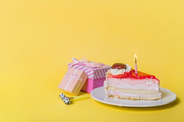 黄色の背景に2つのギフトボックスとパーティーブロワーを持つスライスケーキの照明付きキャンドル