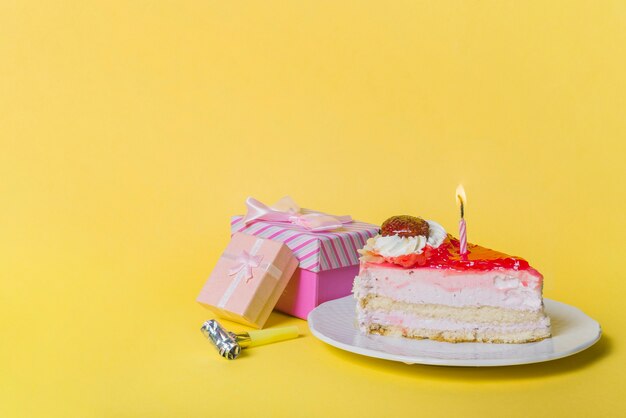 Illuminated candle on slice cake with two gift boxes and party blower against yellow background