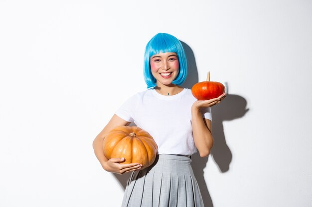 Image of adorable asian girl in blue wig, celebrating halloween, showing large and small pumpkins and smiling happy, standing.