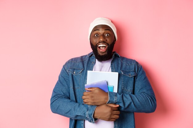 Image of adult african-american man holding notebooks and smiling, studying at courses, standing over pink background