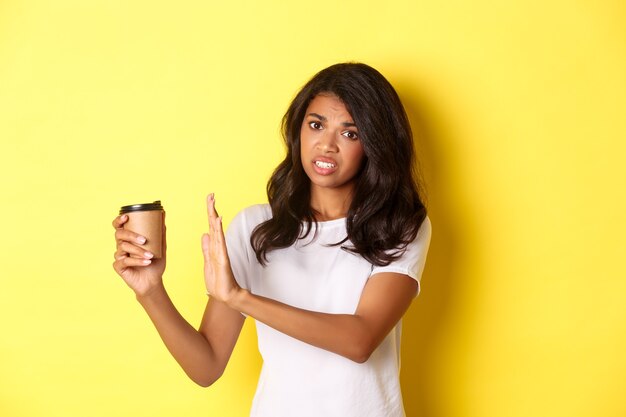 コーヒーの味の悪さを訴え、拒否の兆候を示し、黄色の背景の上に立ってカップを引き離すアフリカ系アメリカ人の女の子の画像。