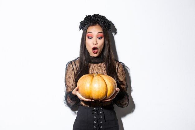 Image of amazed asian girl looking at big pumpkin fascinated, wearing gothic dress and black wreath, standing over white background.