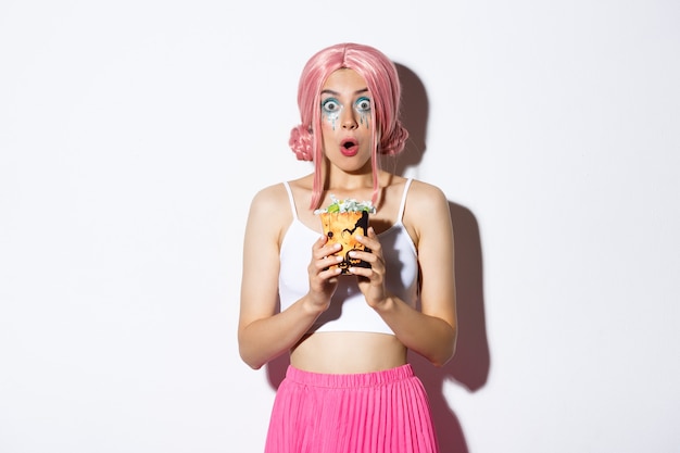 Image of amazed beautiful girl in pink wig, with bright makeup, celebrating halloween, holding candies with surprised face, standing over white background.